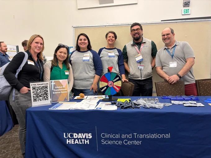 UC Davis Clinical and Translational Science Center team members stand behind an exhibit table at the UC Davis Research Expo with CTSC materials, a QR code sign-up sheet, a prize wheel, and giveaway items.