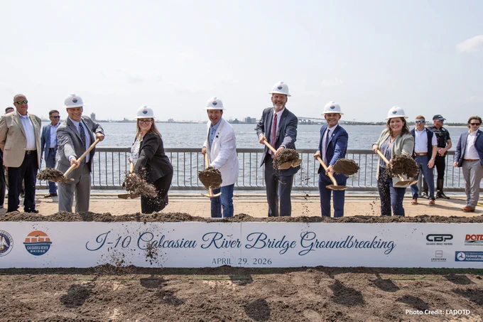 A group of individuals wearing hard hats are participating in a groundbreaking ceremony for the I-10 Calcasieu River Bridge. They are holding shovels with mounds of dirt. A waterfront is visible in the background. The banner indicates the event date as April 29, 2026. Several onlookers are present.

