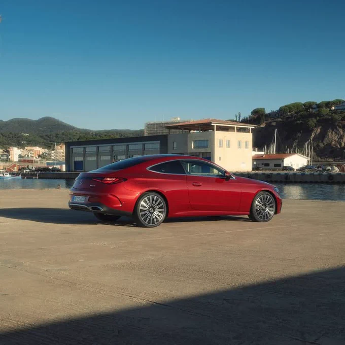 A red sports car is parked on a concrete platform near a waterfront. Buildings are visible in the background under a clear blue sky.