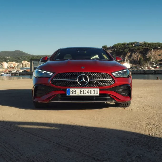 A front-view shot showcases a red Mercedes-Benz with the license plate \"BB EC 4010\", parked on a paved area. In the background, there's a clear blue sky, along with structures that seem to be part of a coastal town or harbor.