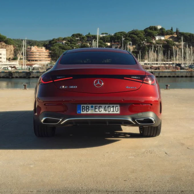 A red Mercedes CLE 300 4MATIC is parked on concrete with a harbor, green hills, and blue sky in the background.