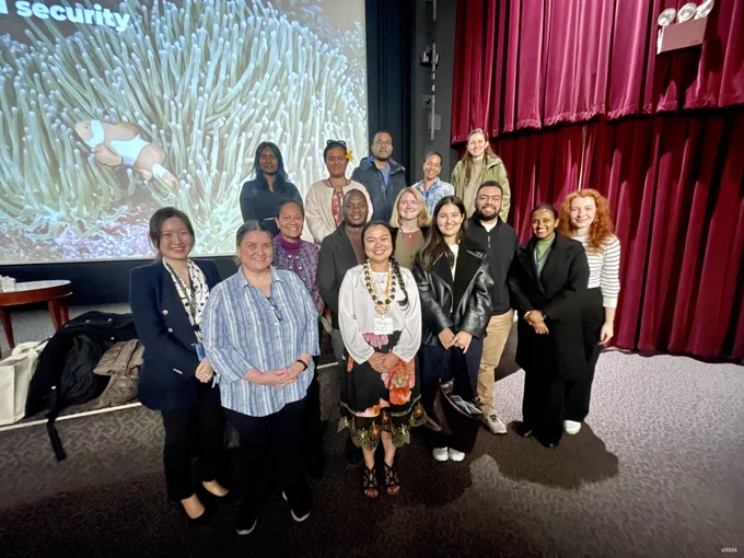 Group of colleagues with marine biologist guest standing in front of a large theater screen displaying coral reef imagery.
