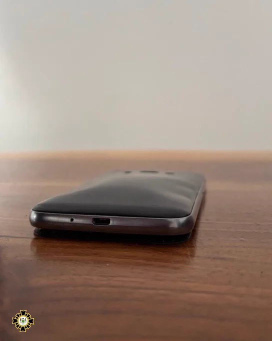 A smartphone with a bulging battery rests on a wooden surface. The phone is positioned face down, showing its bloated back cover. The background is a plain, light-colored wall. The Office of the Fire Marshal logo is in the bottom left corner of the image.