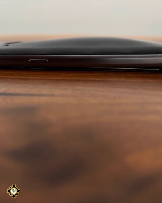A smartphone with a slightly bulging battery lifting the screen from its casing, resting on a wooden surface. The device appears to be damaged due to the battery expansion.