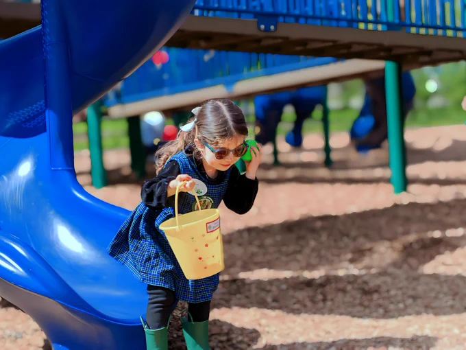 A young girl wearing sunglasses holds a bright green beeping egg up to her ear. She is holding a yellow bucket in a colorful playground.