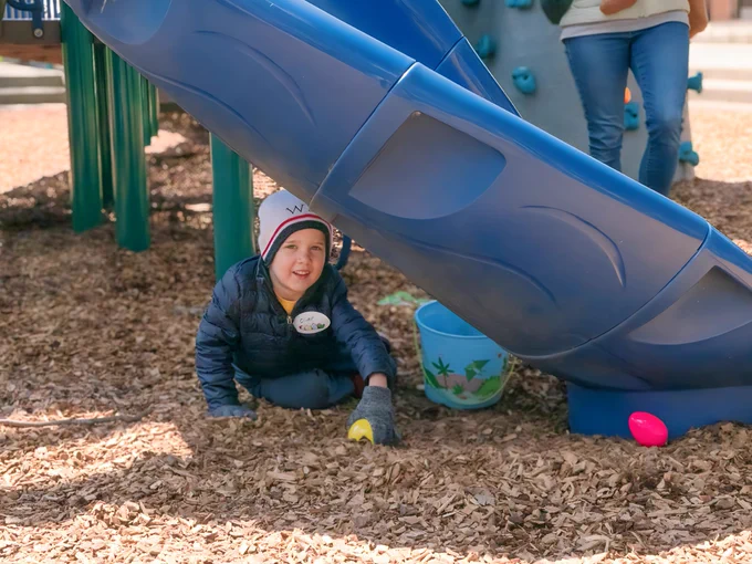 A young boy smiles as he finds two beeping eggs under a blue playground slide. A colorful bucket sits next to him.