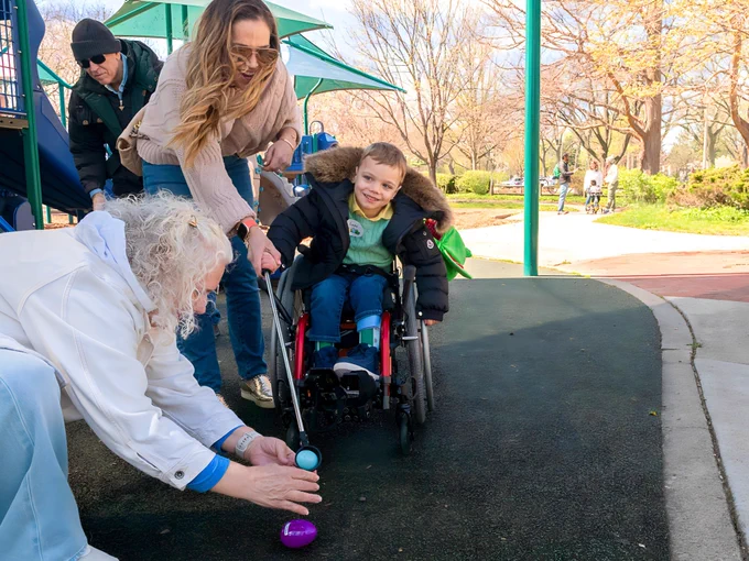 A child in a wheelchair is smiling while participating in an egg hunt at a playground. Adults are assisting, with one placing colored eggs on the ground. 