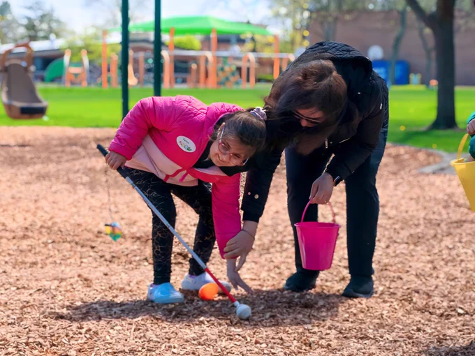 A child in a bright pink jacket reaches down to pick up a beeping egg from the ground . She is holding a white cane and an adult is are in a playground engaging in an Easter egg hunt. The child, holding a white cane, is reaching for an egg on the ground. Both are carrying colorful baskets. The background features playground equipment.