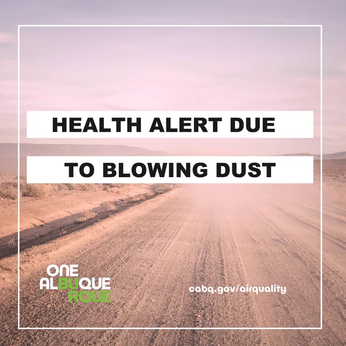 A desert road with dust blowing over it. The text reads health alert due to blowing dust. Green One Albuquerque logo in the bottom corner.