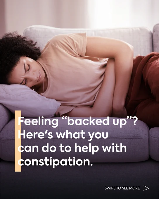 A woman laying on a couch in distress, clutching her stomach. Text reads: \"Feeling 'backed up'? Here's what you can do to help with constipation. SWIPE TO SEE MORE.\"