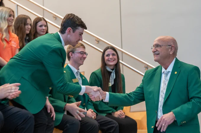 A group of individuals wearing green blazers sits on tiered seating. One person, also in a green blazer, shakes hands with an older person standing at the bottom of the stairs. Others in casual attire are nearby. A railing is visible in the background.