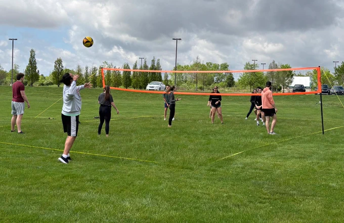 Students compete in an outdoor volleyball match on a grass court.
