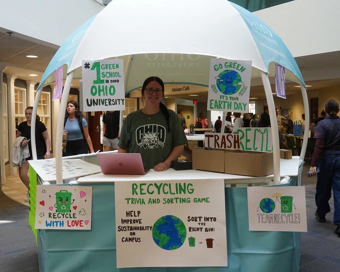 A person stands behind a booth promoting recycling at Ohio University. The booth features green-themed signs and posters with messages such as "Go Green! It's Your Earth Day" and "Recycling Trivia and Sorting Game." There is a laptop and cardboard boxes present.