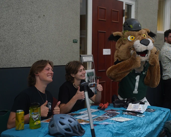 Two people smile and give thumbs up next to OHIO's mascot Rufus the Bobcat.  They are seated at a table covered with brochures and a bicycle helmet. A large water bottle and bike lock are also on the table. 
