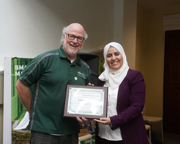 Two individuals stand side by side smiling, with one holding a framed certificate. The person on the left is dressed in a green polo shirt with the "Ohio University" logo. 