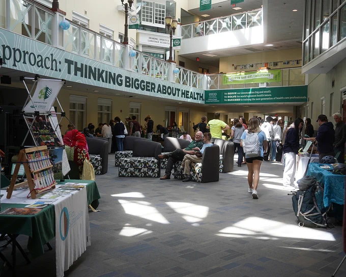 The image shows an indoor event at Ohio University, with attendees mingling in a spacious hall. Several informational booths are set up. The hall is well-lit with natural light from windows, and banners are displayed with messages about health.