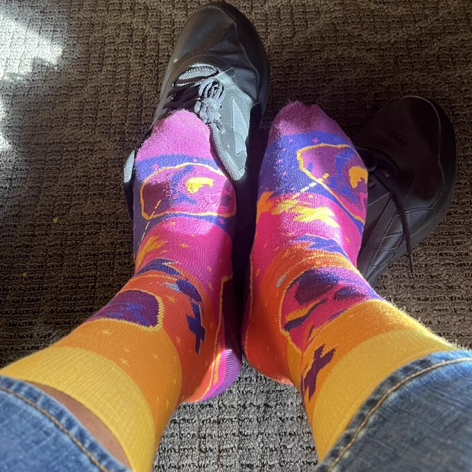Feet wearing Psyche Metal Asteroid Mission socks with a vibrant yellow-to-pink-to-purple gradient and the Psyche spacecraft and 16-Psyche asteroid woven into the design.
