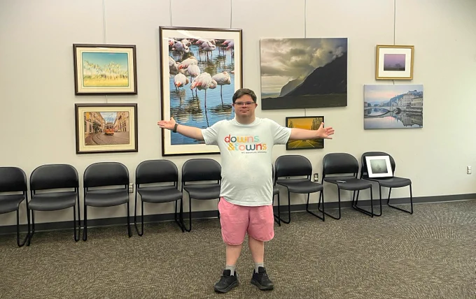 Man with Down syndrome extends his arms in front of his gallery show for his travel photography. The photos behind him are images from France, Iceland, Portugal, and the United States. 