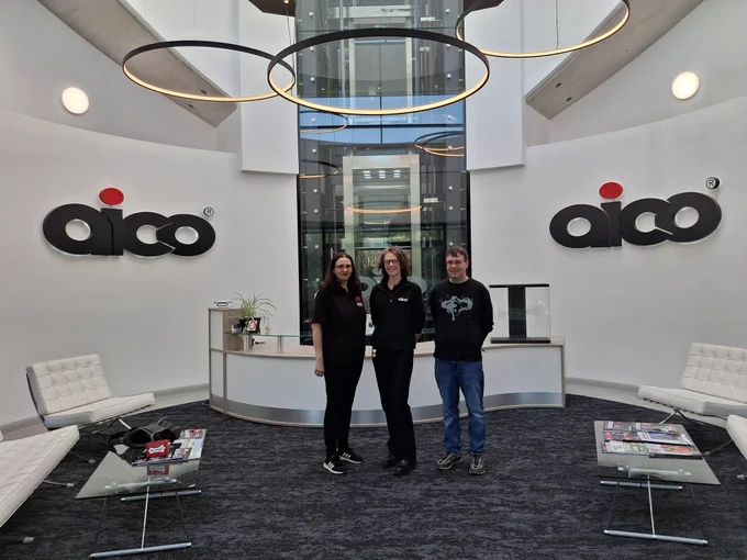 Three people, Rachel, Becci Cumblidge, and Tim stand smiling together in the modern, bright atrium of the Aico building. Rachel and Tim from Red Nought are standing on either side of Becci from Aico. The background features a curved white wall with large "Aico" logos, a glass elevator, and unique circular "halo" light fixtures hanging from the ceiling