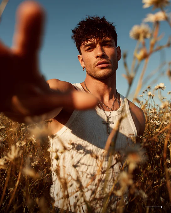 Close-up portrait of a man in a white tank top reaching toward the camera in a sunlit field of wildflowers with warm golden hour lighting.