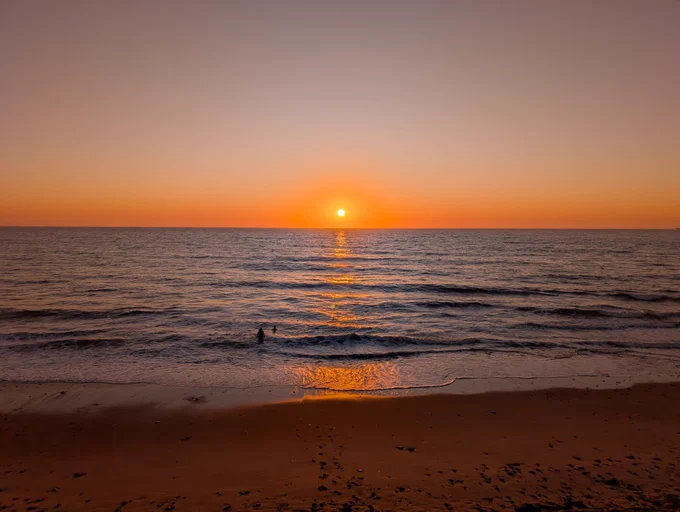 Image shows the sun rising at Whitley Bay on 1st May 2026. Two swimmers can be seen making their way into the sea.