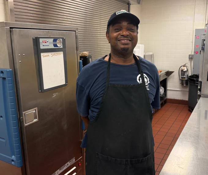 A cafeteria worker wearing glasses and a “Champion” cap holds a small box labeled “School Nutrition Hero Day” in a school cafeteria.