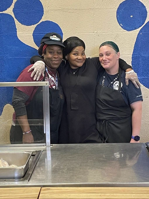 Three cafeteria staff members stand together behind a serving counter, wearing aprons and hair coverings, smiling with their arms around each other.