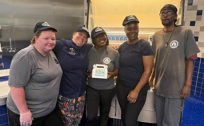Five cafeteria staff members pose in a commercial kitchen; one holds a small white box with a colorful label.