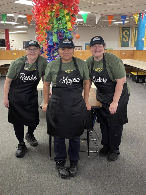 Three cafeteria workers wearing aprons and “Champion” caps stand in a decorated cafeteria with colorful hanging ribbons and banners.