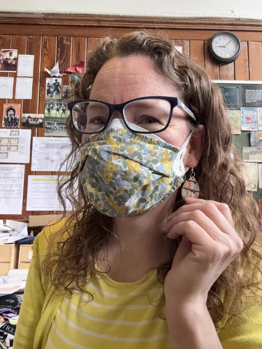 A woman with curly hair and glasses smiles at the camera. She is wearing a yellow and white Breton striped top and a yellow cardigan, along with a green and yellow face mask. Her left hand is showing off an earring in her left ear, which is decorated with books on a shelf.