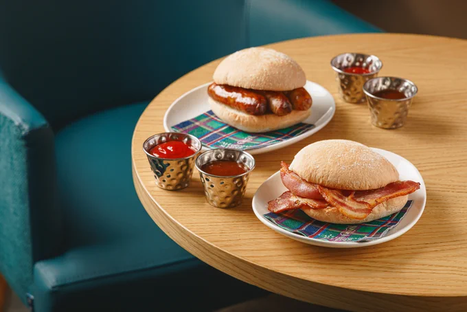 A bacon roll and a sausage roll on round plates with tartan napkins with sauce on a round table.