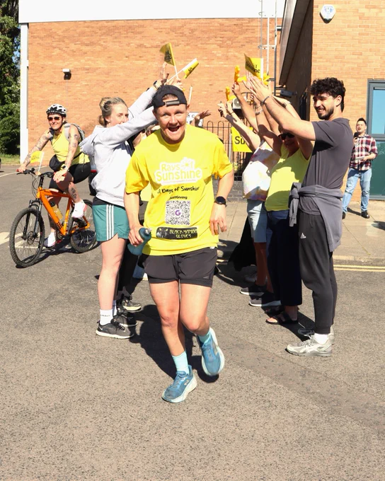 Smiling runner in yellow charity shirt passes through cheering crowd forming a raised-hands tunnel.