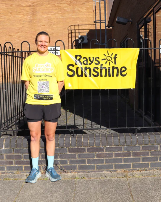 Smiling woman in a yellow "Rays of Sunshine" shirt standing beside a yellow "Rays of Sunshine" banner hung on a black metal fence
