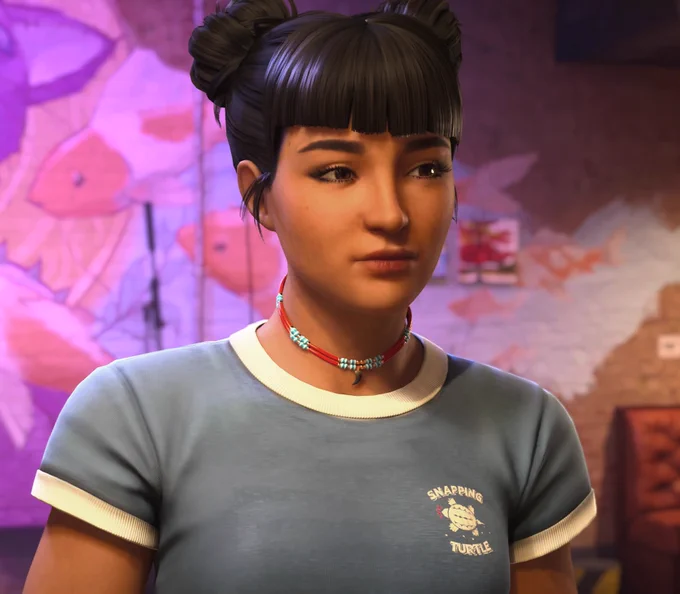 Amanda Thomas from Life is Strange: Double Exposure, an Indigenous woman of Cherokee and Algonquin descent, posing in her bar, The Snapping Turtle. She has dark hair styled in space buns and blunt bangs, and is wearing high-waisted jeans and a light blue baby tee with the bar's logo. The warm interior of the Lakeport, Vermont bar is visible in the background.