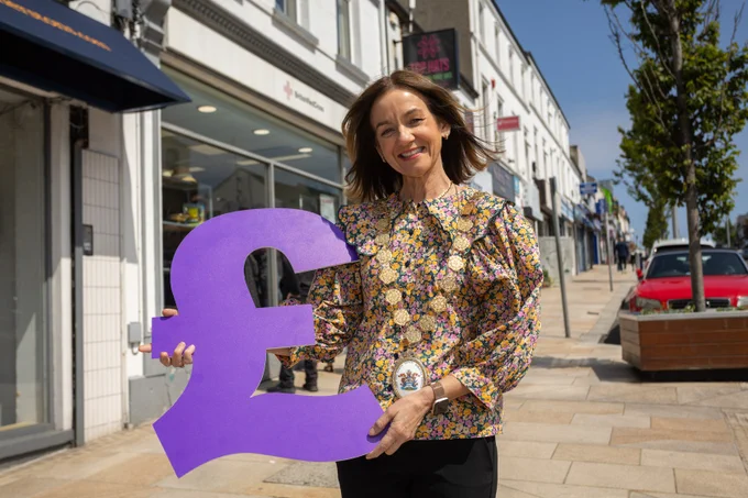 Mayor Councillor Gillian McCollum is holding a large purple British pound symbol on a sunny street lined with shops and parked cars.