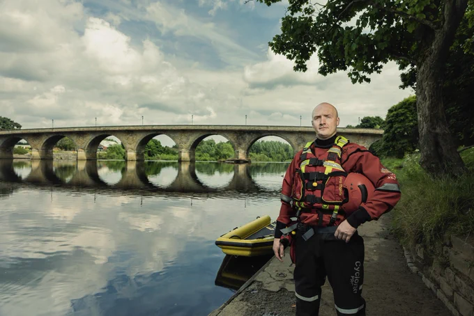 Firefighter Sean, wearing water rescue gear standing by Hexham Bridge with a water rescue floatation raft in the river beside him