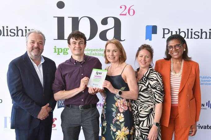 The image is a photograph taken at the Independent Publishers Guild Awards 2026 featuring 5 people. The background features the IPA logo and in front of it are 5 people, with the middle two holding the Virtusales Metadata Award. From L-R, the people are: Hal Cruttenden, Alex Harrison (Bloomsbury), Fiona Green (Bloomsbury), Kat Coveyduck (Virtusales), and Juliette Foster.