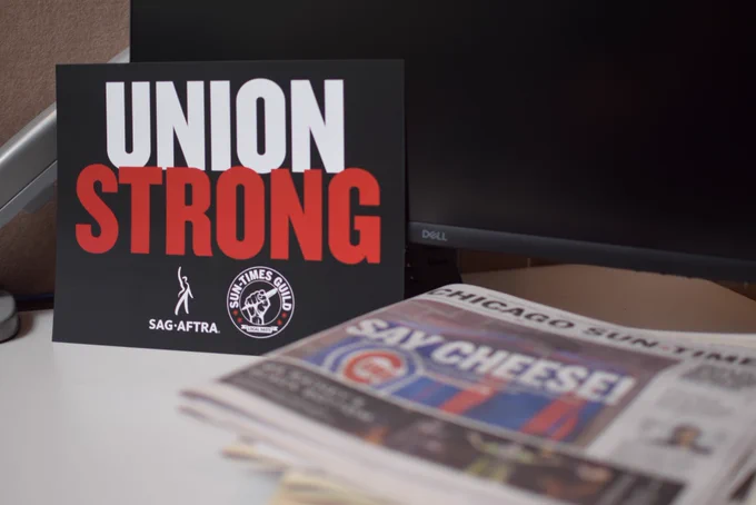 A UNION STRONG sign with both the guild and WBEZ SAG-AFTRA logos on it sits next to a copy of the Sun-Times paper with the Wrigley ray on the cover.