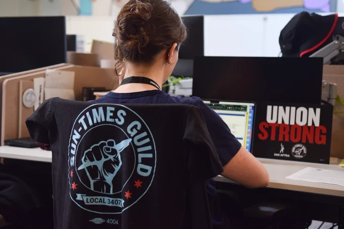 Sun-Times reporter Kaitlin Washburn works at her desk with a Sun-Times Guild t-shirt on her chair and a UNION STRONG sign with both the guild and WBEZ SAG-AFTRA logos on it.
