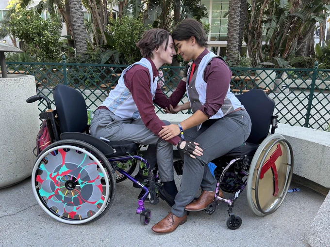 Two wheelchair users dressed as Jayce and Viktor from Arcane scoot forward in their chairs to touch foreheads lovingly. 