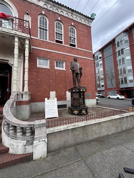 Picture of the right side a red brick, two story building that is the Chong Wa Benevolent Association building in Chinatown Seattle, WA. On the left side, part of the front door shows with a concrete handrail leading up to the door that has two large, grey columns to the right of the door. On top of the door there is a balcony that is on the second story. To the right of the front door is a statue of Dr. Sun Yat-sen on top of a large, black pedestal with gold writing in Chinese. 