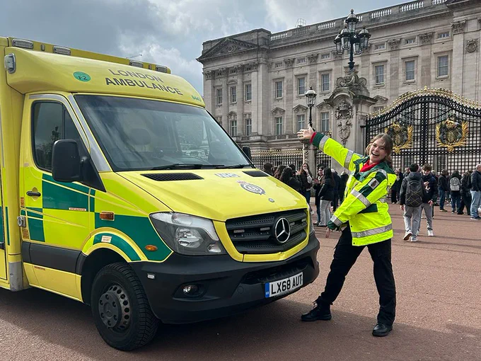 An ambulance is parked in front of Buckingham Palace. A person in a high-visibility jacket is standing beside the ambulance, gesturing towards it. Crowds are gathered near the palace gates.
