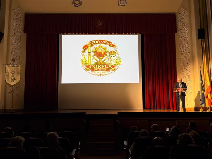 Logotipo "Corpus Valencia 700 años" proyectado en pantalla de teatro; orador en podio a la derecha y público en penumbra.
