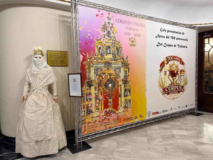 Moma, vestida de blanco y velo, junto a carteles del 700 aniversario del Corpus Christi de Valencia (1326-2026)