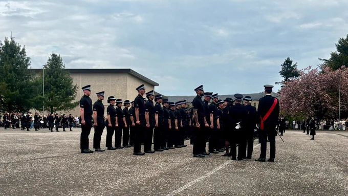 Police officers in uniform standing in formation outdoors, with a pipe band visible in the background.