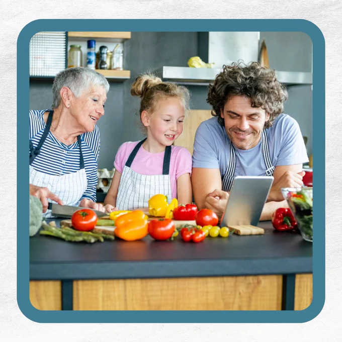 Three people in a kitchen, smiling and looking at a tablet, with colorful vegetables on the countertop.