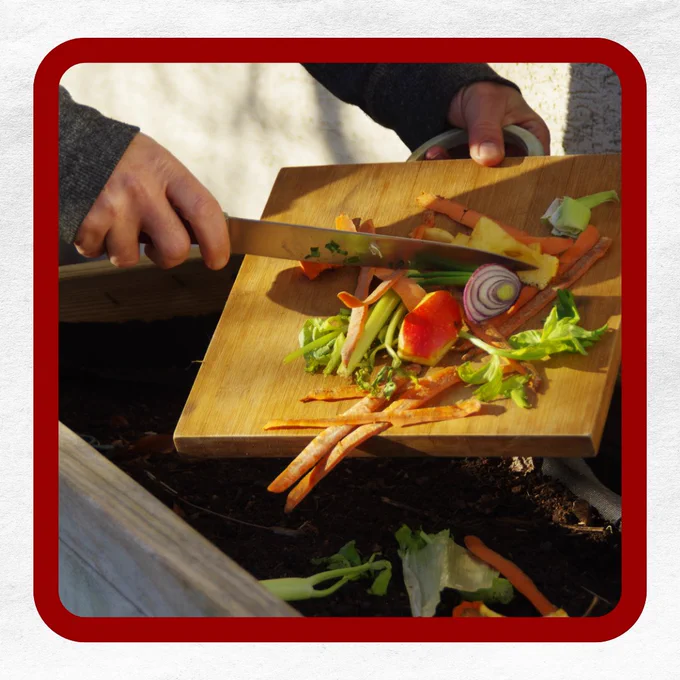 Person scraping vegetable scraps into a compost bin using a wooden cutting board.