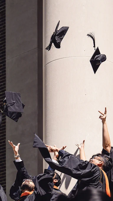 Graduates in caps and gowns celebrating by throwing their hats in the air in front of a building with large columns. "MAY" is written in the top left corner.
