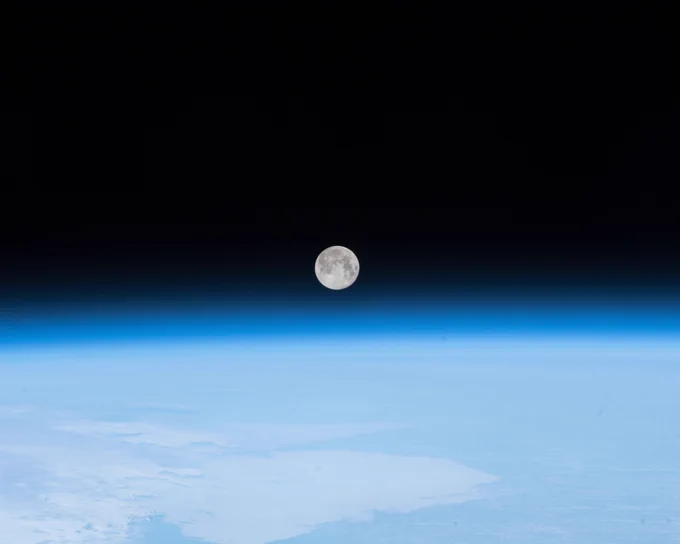 A full moon is centered in the image, against the inky black of space. Below, a blue haze from Earth's atmosphere glows, while clouds, land, and oceans cover the rest of the planet in view. Credit: NASA