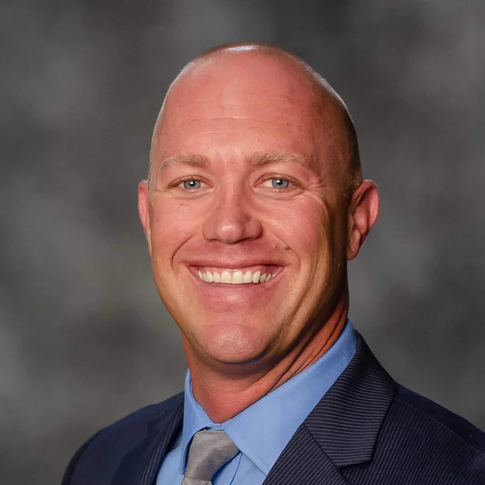 Smiling bald man in a blue suit and tie, against a blurred gray background.
